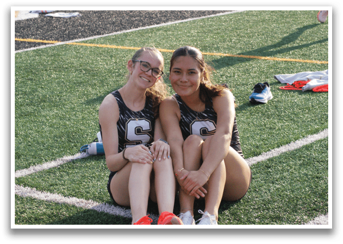 Two female athletes wearing uniforms sit on the grass. Contenido generado con IA