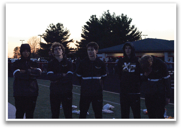 Five young men wearing black jackets and hoods stand on a field. Contenido generado con IA