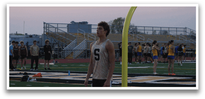 A man stands on a track field, wearing a jersey and shorts. He is the only person on the field. Contenido generado con IA