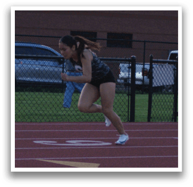 A woman is running on a track, wearing a black shirt and white shoes. She is running in front of a building. There are several cars parked in the background. Contenido generado con IA