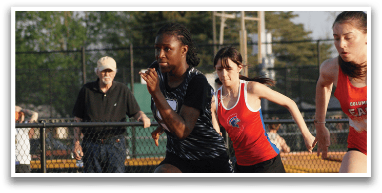 Two girls are running on a track, one in front of the other. They are wearing shorts and are in motion. A man is watching them from behind a fence. Contenido generado con IA