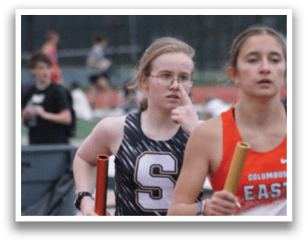 Two girls are running on a track, one wearing a black and white outfit and the other wearing an orange and white outfit. They are holding their respective batons and appear to be competing in a race. Contenido generado con IA