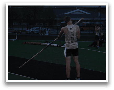 A man in a white shirt and black shorts is holding a pole and standing on a track. Contenido generado con IA