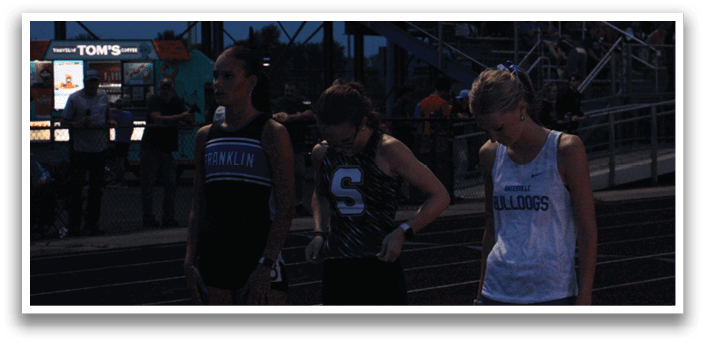 Three female athletes stand on a track, wearing uniforms and waiting for the race to begin. Contenido generado con IA