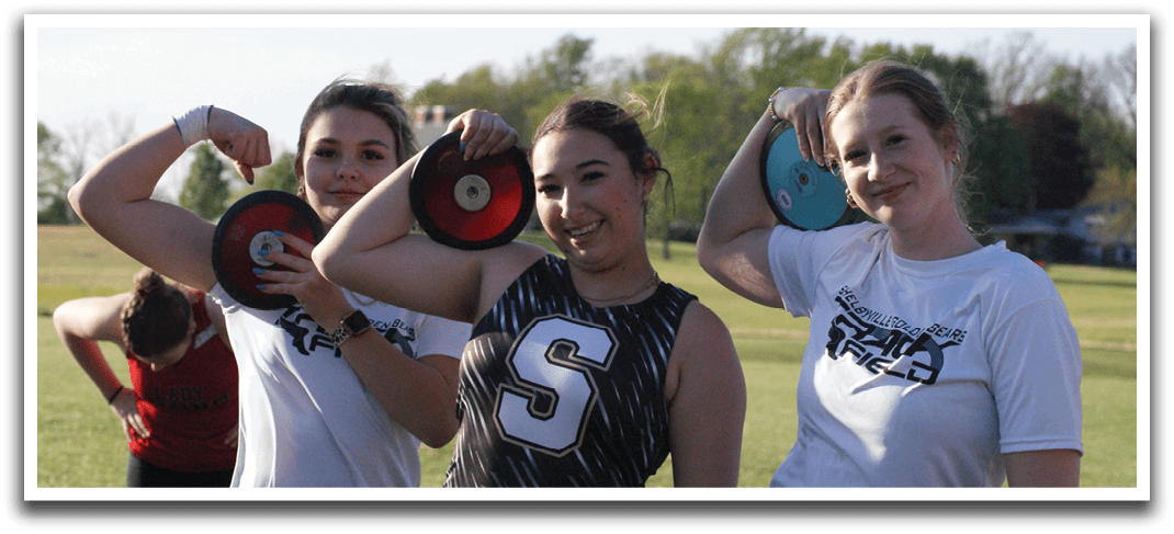 Three women are standing in a grassy field, holding frisbees. They are posing for a picture, with one woman on the left, another in the middle, and the third on the right. The women are wearing shorts, and they are all smiling for the camera. Contenido generado con IA
