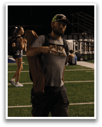 A man wearing a backpack and a hat is standing on a field with other people. Contenido generado con IA