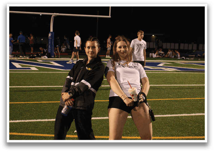 Two women pose on a football field, one with a knee brace and the other with a water bottle. Contenido generado con IA