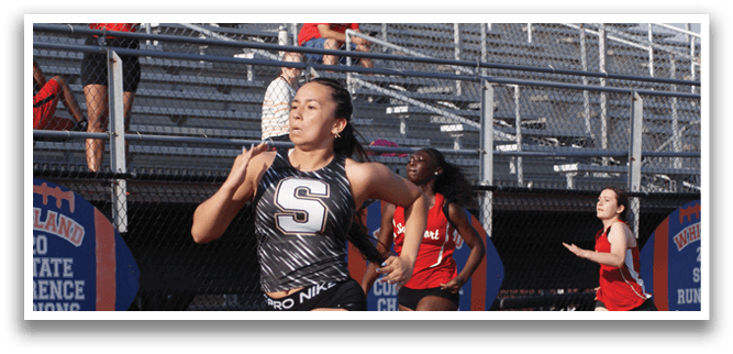 A female athlete is running on a track, wearing a black and white outfit. She is in the lead, with other runners behind her. The scene takes place in a stadium, with spectators watching the event. Contenido generado con IA