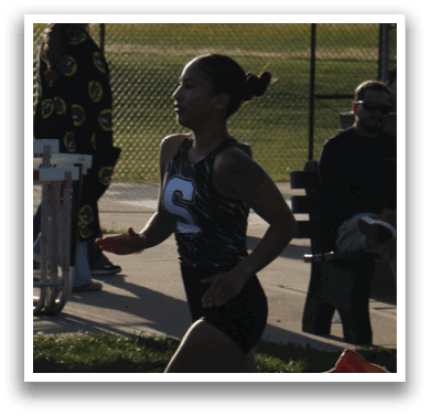 A woman is running on a track, wearing a black and white shirt. She is in the lead and appears to be in the middle of a race. There are several people watching her from the sidelines, including a man and a woman sitting on a bench. Contenido generado con IA