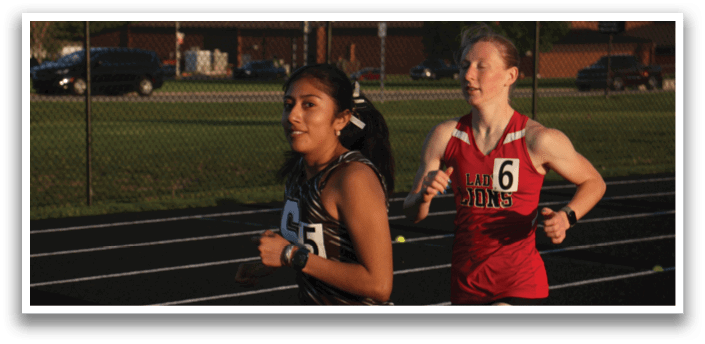 Two female runners are on a track, one wearing a black and white shirt and the other wearing a red shirt. They are both running in the same direction. Contenido generado con IA