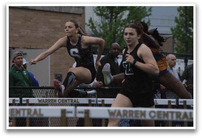 Two female athletes are running on a track, with one of them wearing a black shirt and the other wearing a green shirt. They are both running towards a barrier, possibly in a competition. There are several other people in the scene, some of whom are also running, while others are watching the event. Contenido generado con IA