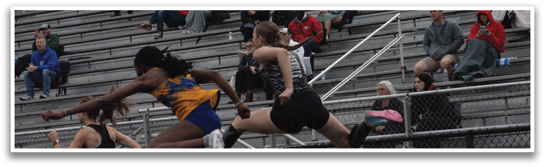 Two female athletes are jumping over a hurdle in a stadium, with spectators watching from the stands. Contenido generado con IA