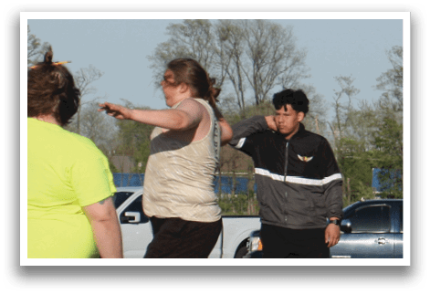 A group of people are standing on a baseball field, with some of them wearing baseball gloves. They are gathered around a woman who is throwing a ball. There is a truck parked nearby. Contenido generado con IA