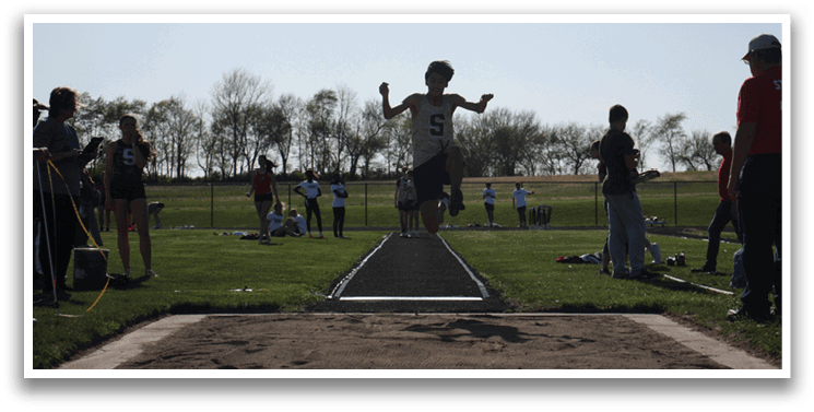 A man is jumping in the air on a track, with several people watching. Contenido generado con IA