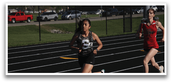 Two girls are running on a track, one wearing a red shirt and the other wearing a black shirt. They are both running fast, with the girl in the black shirt slightly ahead of the girl in the red shirt. The track is surrounded by cars parked in the background. Contenido generado con IA