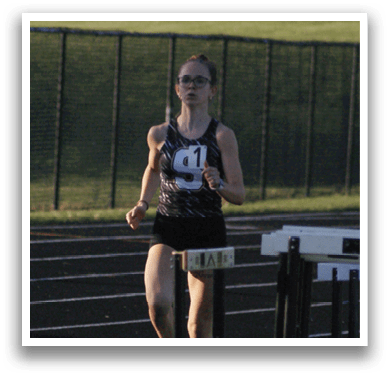 A woman is running on a track, wearing a black shirt and shorts. She is running past a set of hurdles. Contenido generado con IA