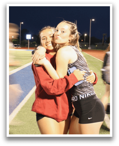 Two women hugging on a track field, one wearing a red shirt. Contenido generado con IA