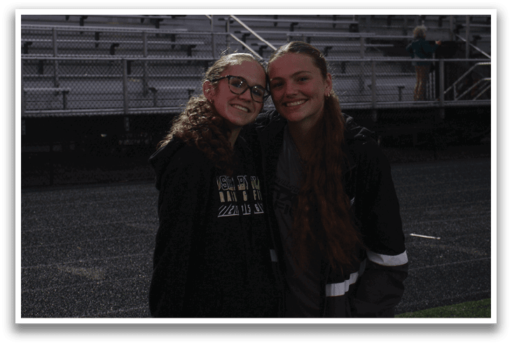 Two girls wearing glasses and hoodies pose for a picture on a field. Contenido generado con IA