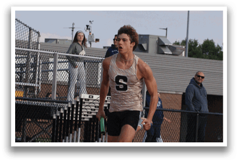 Two men are running on a track, one wearing a blue shirt and the other wearing a white shirt. They are both holding a baton. Contenido generado con IA