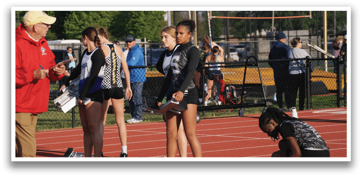 A group of female athletes are standing on a track, some of them wearing black and white uniforms. They are talking to a man, who is likely a coach or an official. The scene takes place in a park, with cars parked in the background. Contenido generado con IA