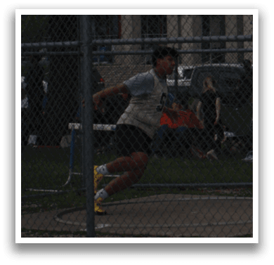 A man is running in a fenced-in area, with a crowd of people watching. There are several cars parked nearby, and a baseball bat is visible in the scene. Contenido generado con IA