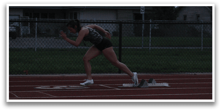 A woman is running on a track, wearing a black shirt and black shorts. She is running in front of a fence and a building. Contenido generado con IA