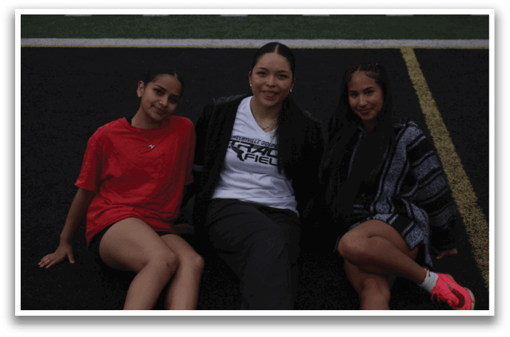 Three people are sitting on a field, two women and a man. The man is wearing a white shirt and the women are wearing black. Contenido generado con IA