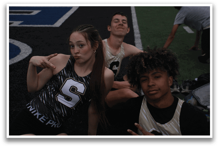 Three young people are sitting on the ground, posing for a picture. One of them is wearing a track suit and making a peace sign. Contenido generado con IA