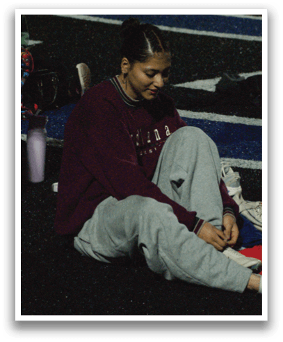 A woman is sitting on the ground with her legs crossed, wearing a maroon sweatshirt and gray sweatpants. She is holding a shoe in her hand. Contenido generado con IA