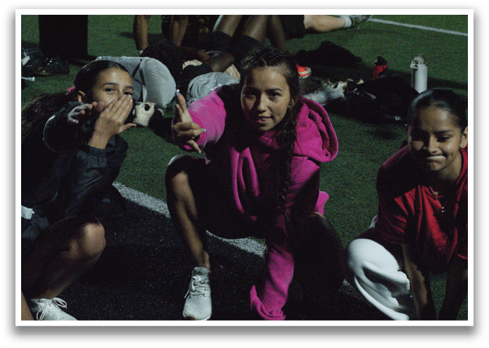 Three girls are sitting on the ground, posing for a picture. One girl is pointing at the camera, while the other two are sitting behind her. They are all wearing pink jackets. Contenido generado con IA