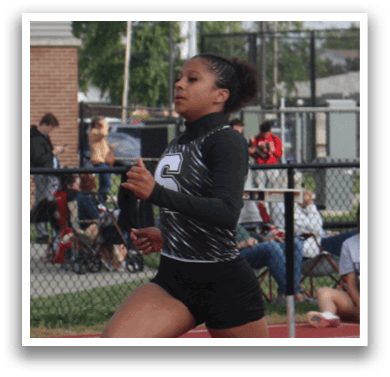 A woman is running on a track, wearing a black shirt and shorts. She is in the lead, with other runners behind her. There are several people in the background, some of them carrying backpacks. Contenido generado con IA