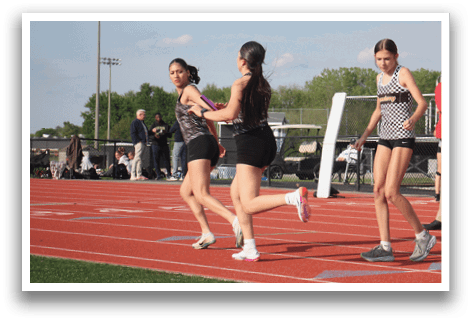 Three female runners are on a track, wearing shorts and running shoes. They are running in a line, with one runner slightly ahead of the other two. Contenido generado con IA