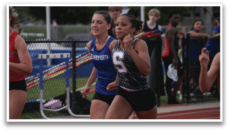 Three female runners are competing in a race. One runner is wearing a blue shirt, another is wearing a red shirt, and the third is wearing a black and white shirt. They are all running on a track. Contenido generado con IA
