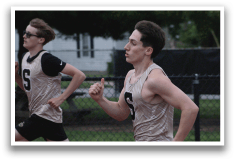 Two young men are running on a track, wearing shorts and shirts. One of them is wearing a number 5 shirt. Contenido generado con IA