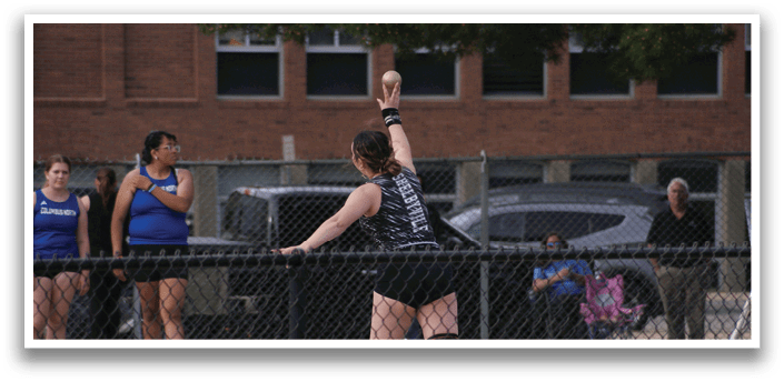A woman is throwing a ball in a fenced-in area, with a crowd of people watching her. Contenido generado con IA