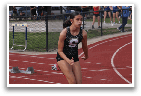 A female athlete is running on a track, wearing a black and white uniform. She is in the lead, with other runners behind her. There are several cars parked in the background, and a bench is visible in the foreground. Contenido generado con IA