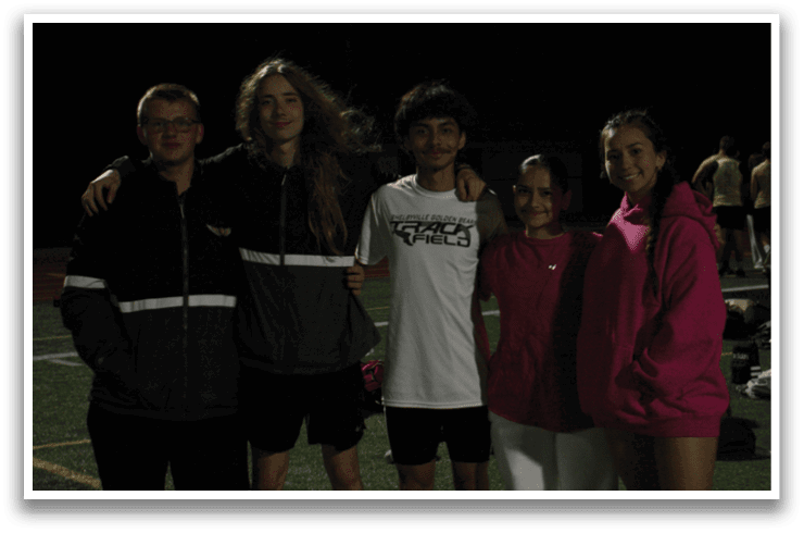 Four people are standing on a field at night, posing for a picture. They are wearing sports uniforms and are surrounded by a pile of clothes. Contenido generado con IA