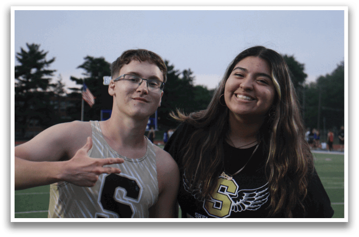 A man and woman pose for a picture, both wearing glasses. The man is wearing a gold and black track suit and the woman is wearing a black shirt. Contenido generado con IA
