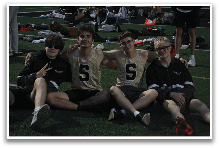 Four young men are sitting on the grass, posing for a picture. They are wearing sports uniforms and are likely teammates. Contenido generado con IA