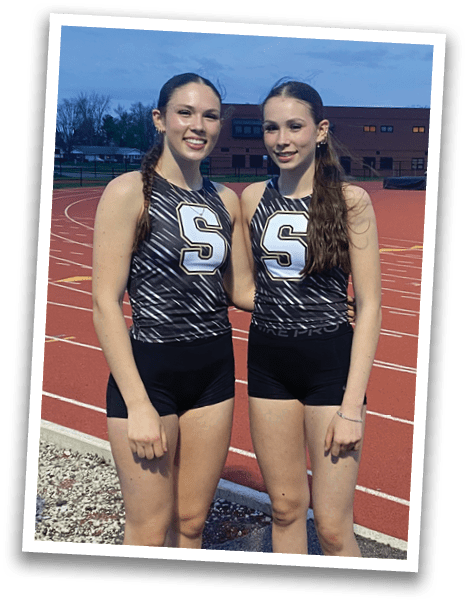 Two female runners in uniforms pose for a picture on a track. Contenido generado con IA