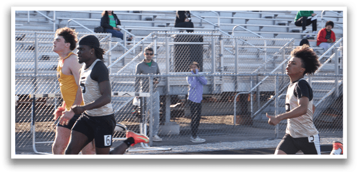 Two men are running on a track, with one of them wearing a black shirt and the other wearing a yellow shirt. They are both running towards the finish line. There are several people watching the race from the stands, some of them sitting on chairs and others standing. The spectators are spread out across the stands, with some closer to the front and others further back. Contenido generado con IA
