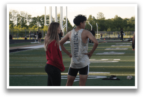 A man and woman are standing on a football field. The man is wearing a tank top and the woman is wearing a red shirt. Contenido generado con IA