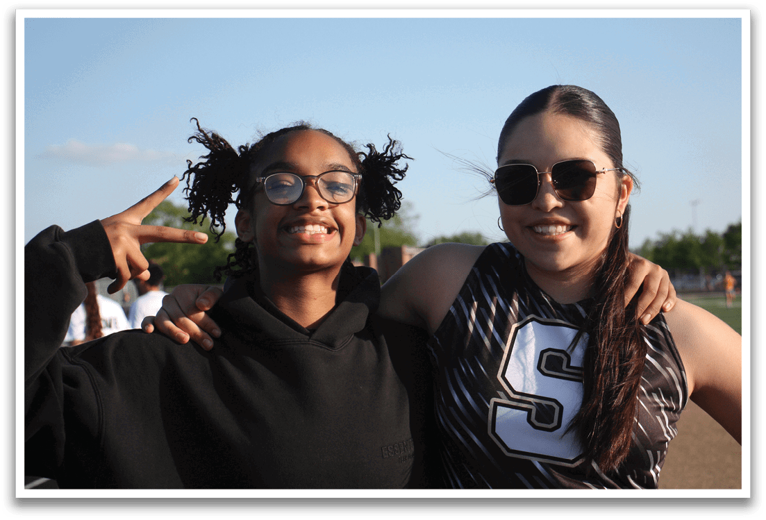 Two young women are standing together, one wearing a black hoodie and the other wearing a black and white outfit. They are both smiling and posing for a picture. Contenido generado con IA
