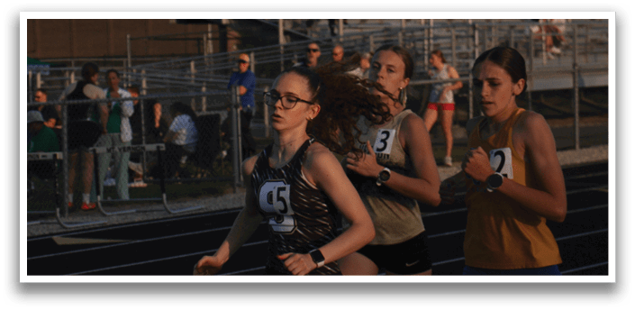 Three female runners are racing on a track, with one of them wearing a black and gold shirt. They are running in front of a crowd of spectators. Contenido generado con IA