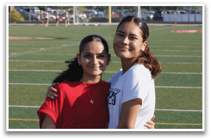 Two girls wearing red and white shirts are standing on a field. Contenido generado con IA