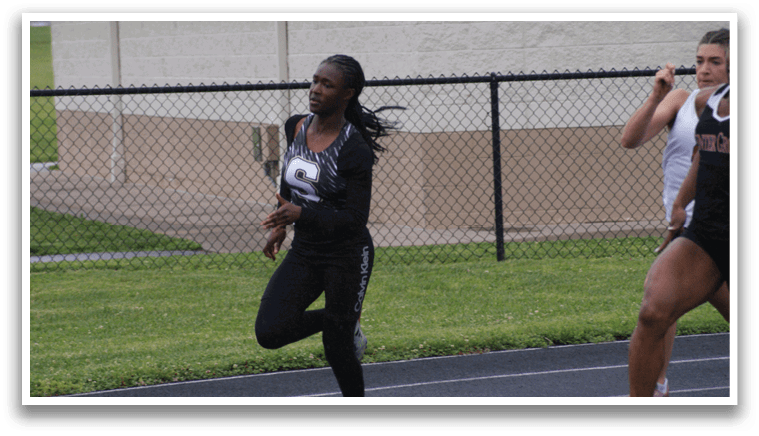 Two women are running on a track, one in front of the other. The woman in front is wearing a black track suit and appears to be running faster than the other woman. Contenido generado con IA