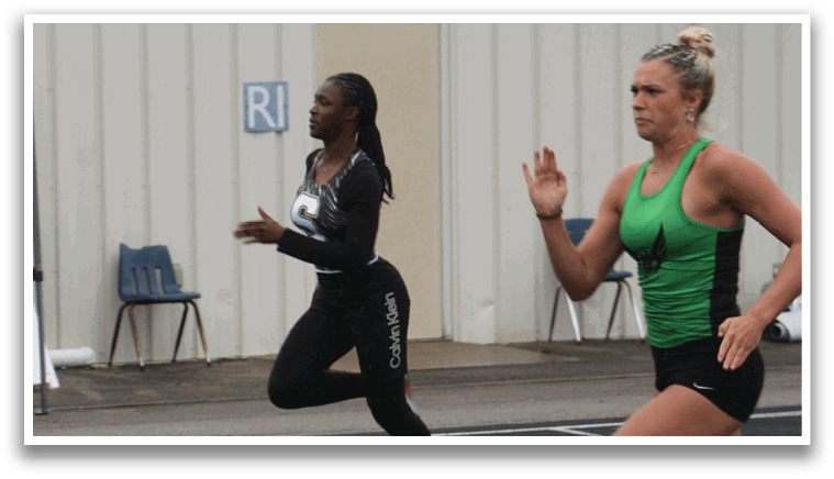Three women are running on a track, with one wearing a green shirt and the other two wearing black. They are all running in the same direction, with one of the women in the lead. Contenido generado con IA