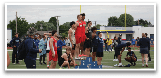 A group of people are standing on a field, with some of them on a blue box. A man is standing on a box, possibly judging an event. There are several people in the background, and a few cars are parked nearby. Contenido generado con IA