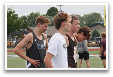 A group of men are standing on a track, some wearing shorts and others wearing shirts. They are lined up on a blue mat, possibly for a competition. Contenido generado con IA