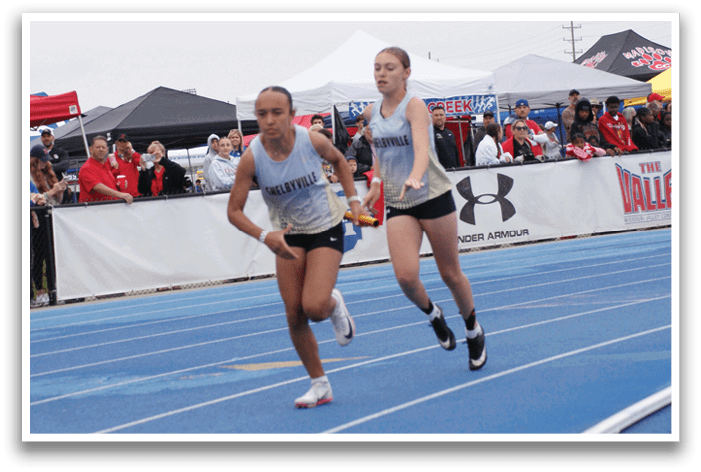 Two female runners are competing in a race, with one of them wearing a blue shirt. They are running on a track in front of a crowd of spectators. Contenido generado con IA